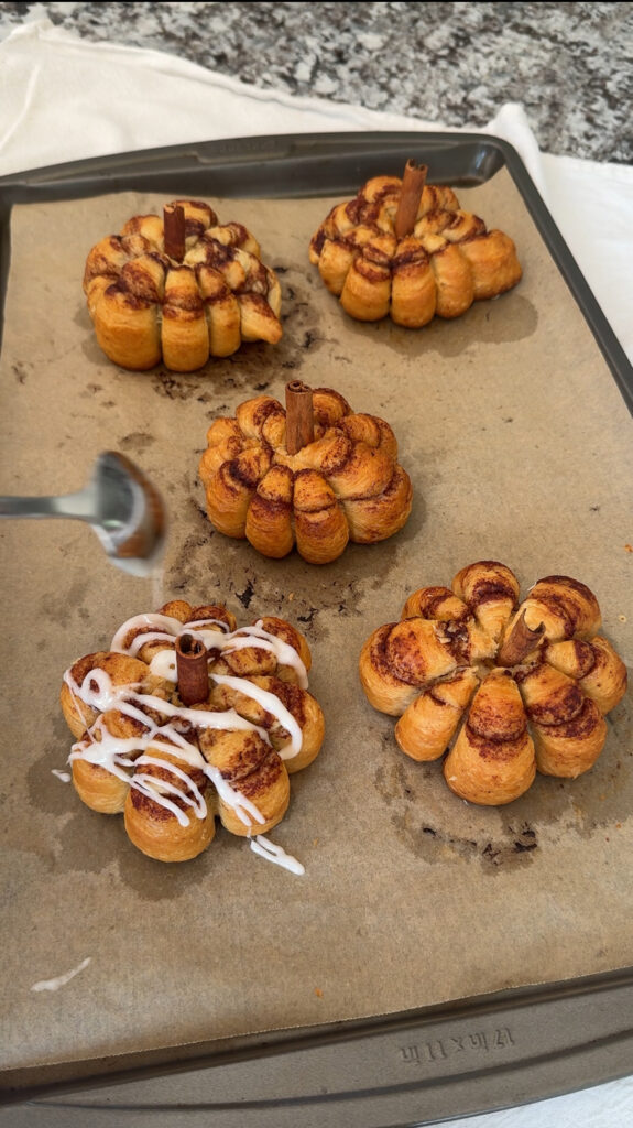 Cooked cinnamon rolls with a cinnamon stick stem are drizzled with icing.